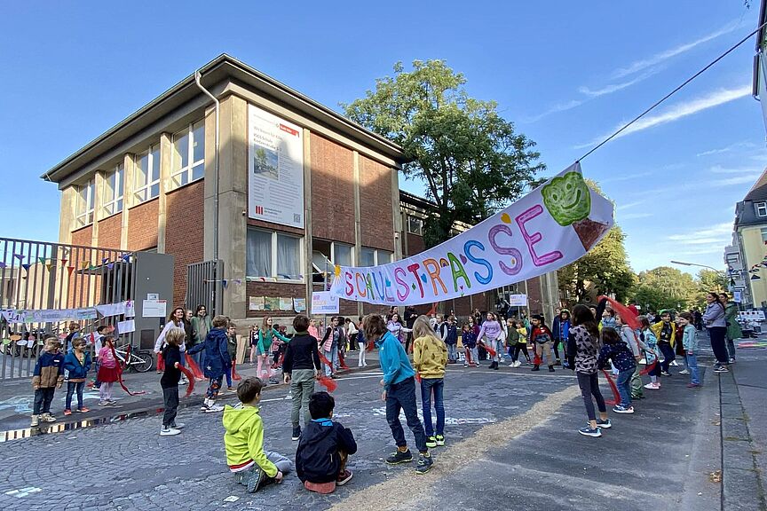 Schulstraße Kinder verschiedenen Alters spielen und bewegen sich auf einer gepflasterten Straße vor einem zweistöckigen Backsteingebäude. Über der Straße hängt ein buntes Banner mit der Aufschrift „Schulstraße“ und einem grünen Baum-Symbol. Im Hintergrund sind Erwachsene zu sehen, die die Aktion begleiten.